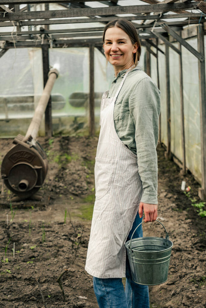 An attractive woman holding a tin bucket and walking in the greenhouse looking at seedlings  - Photo, Image