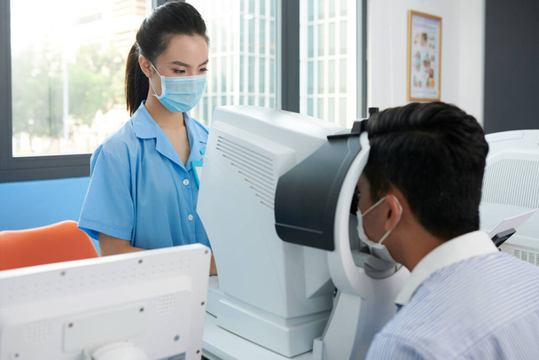 Молода офтальмологічна медсестра в медичній масці перевіряє очі пацієнта на різні захворювання
 - Фото, зображення