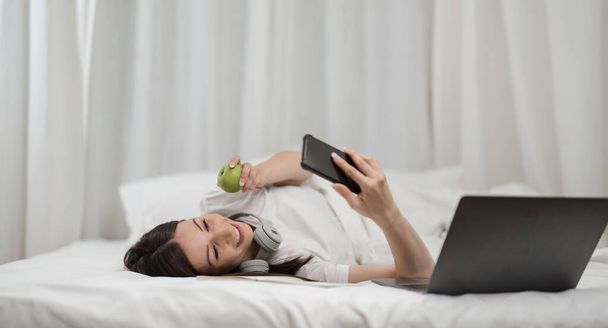 Молодая женщина расслабляется и едят фрукты с помощью телефона на проверку социальных приложений и работы в спальне. - Фото, изображение