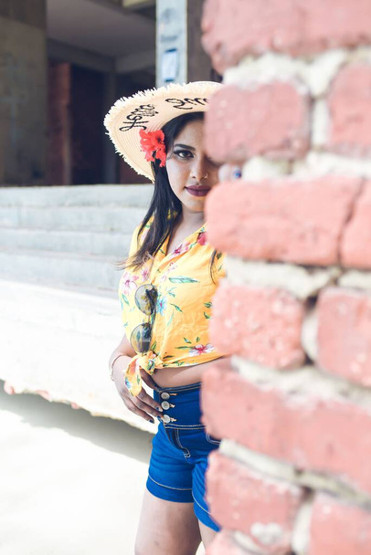 A vertical of a South Asian female in a floral yellow shirt and jeans posing against a brick wall - Fotó, kép