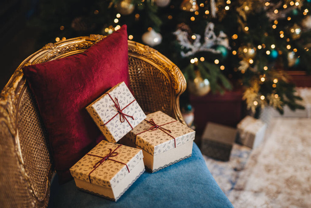 Three wrapped present boxes on armchair near Christmas fir tree. Design or idea for postcard. New Year composition with decorated fir tree and gifts. Majestic atmosphere. Festive event - Zdjęcie, obraz