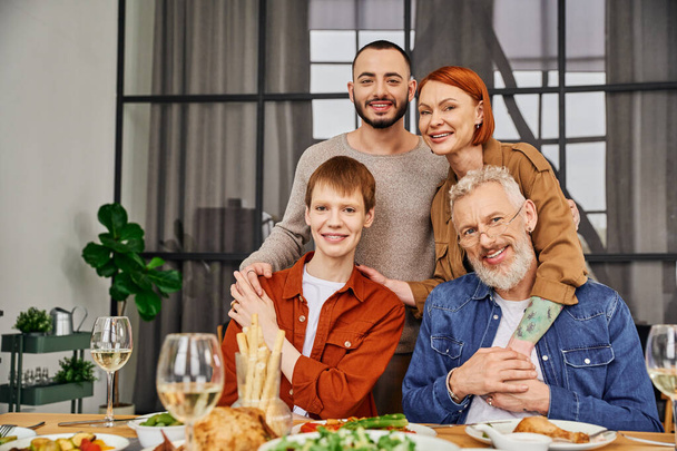 young and happy gay couple with smiling parents embracing and looking at camera near supper served in living room - Photo, Image