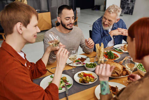 Χαρούμενος γενειοφόρος γκέι άντρας που μιλάει κοντά σε φίλο και γονείς κατά τη διάρκεια του οικογενειακού δείπνου στο σπίτι - Φωτογραφία, εικόνα