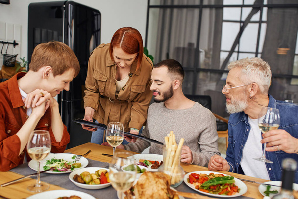 молодые партнеры-геи смотрят фотоальбом во время вкусного семейного ужина на кухне - Фото, изображение