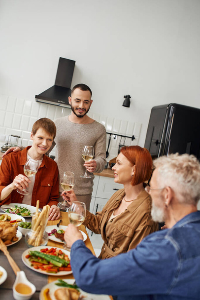 young gay man toasting with wine during supper with boyfriend and happy parents in kitchen - Photo, Image