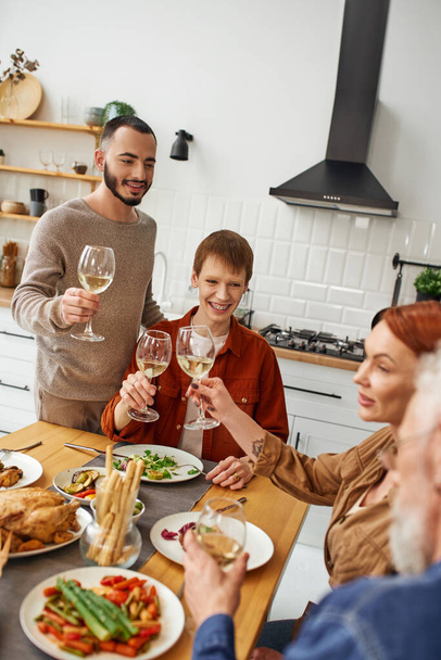 young gay couple with happy parents toasting with wine during family supper in kitchen - 写真・画像