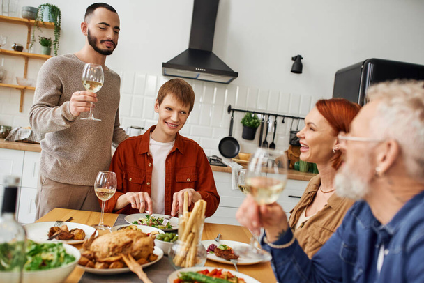bärtiger schwuler Mann stößt mit Wein in der Nähe von Freund und glücklichen Eltern beim Familienessen in der Küche an - Foto, Bild