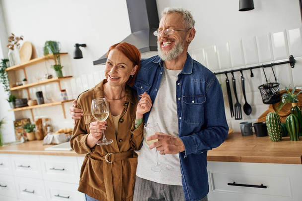 happy redhead woman holding wine glass and smiling at camera near excited husband laughing in kitchen - Photo, Image