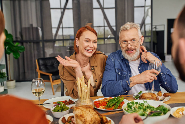 complacidos padres sonriendo cerca borrosa pareja gay durante la cena familiar en sala de estar - Foto, Imagen