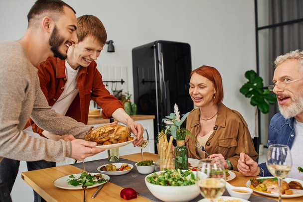 joyful gay couple serving grilled chicken near happy parents in kitchen - Foto, immagini