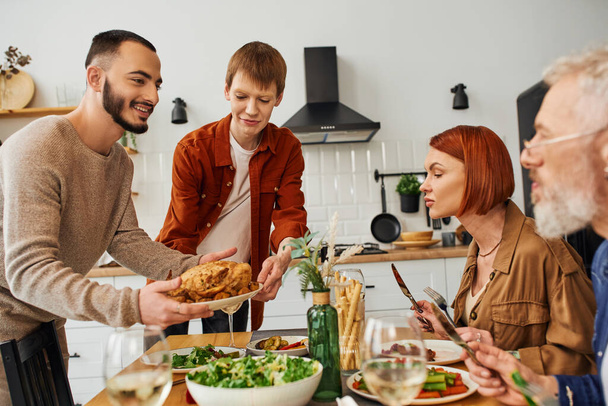 cheerful gay couple serving grilled chicken for family supper with parents in kitchen - Photo, Image