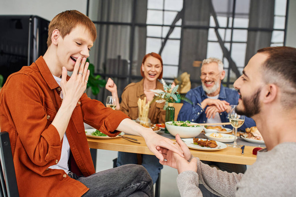 sorprendido hombre cubriendo la boca mientras novio haciendo matrimonio propuesta cerca feliz padres durante la cena familiar en sala de estar - Foto, imagen