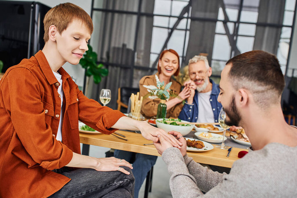 бородатий гей чоловік у обручці на щасливому хлопці біля смачної вечері та щасливих батьків у вітальні
 - Фото, зображення