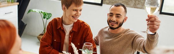 bearded gay man toasting with wine near smiling boyfriend and blurred mother in kitchen, banner - 写真・画像