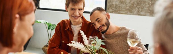 ruda człowiek patrząc na rozmyte rodzice w pobliżu zadowolony gej partner i wino okulary w domu banner - Zdjęcie, obraz
