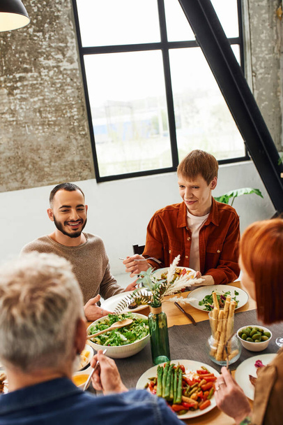 Рыжий гей улыбается рядом с бородатым парнем и размытыми родителями с вкусной едой, подаваемой на кухне - Фото, изображение