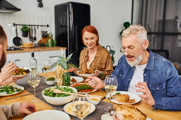cheerful bearded man in eyeglasses talking to family while having delicious dinner in kitchen - 写真・画像