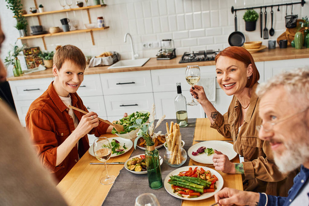 happy gay man with parents looking at blurred boyfriend while having delicious supper in kitchen - Photo, Image