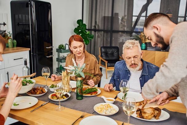 bearded gay man cutting grilled chicken during family dinner with boyfriend and parents in kitchen - Photo, Image