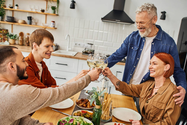 young gay man clinking wine glasses with boyfriend and happy parents during family supper in kitchen - Zdjęcie, obraz