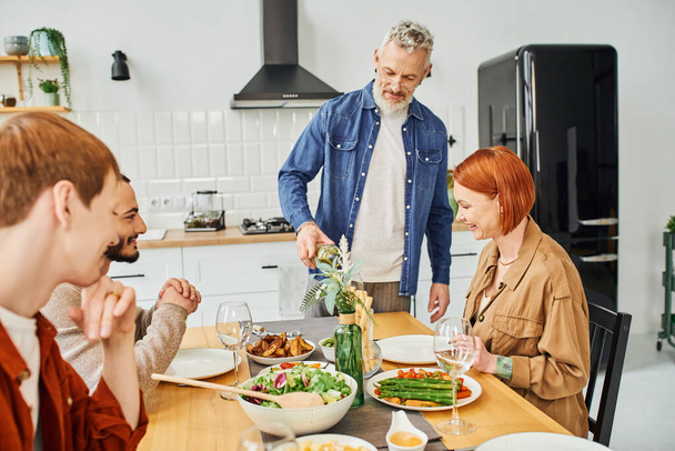 бородатый человек наливает вино рядом с счастливой семьей, обедая вкусно на современной кухне - Фото, изображение