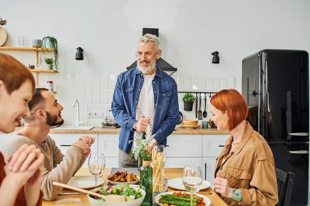 sonriente barbudo hombre apertura vino botella cerca familia cena con hijo y gay hombre en cocina - Foto, imagen