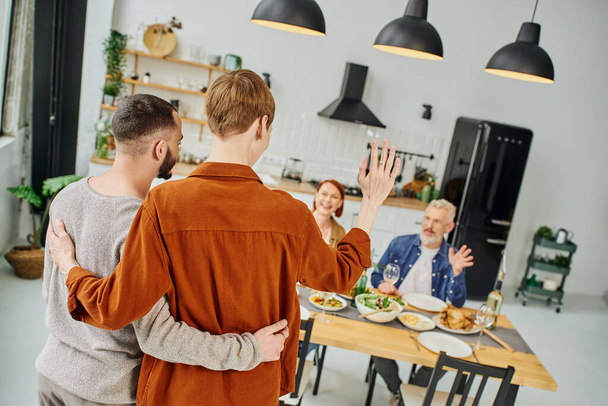 rousse gay homme embrassant copain et agitant la main pour parents ayant souper dans la cuisine - Photo, image