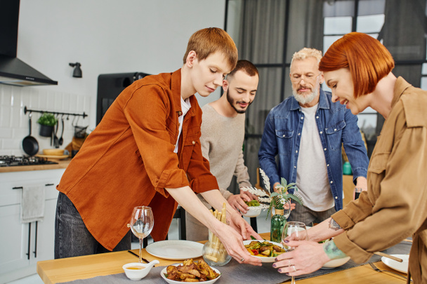 redhead gay man with mother serving grilled vegetables on table with delicious supper in kitchen - Foto, Bild