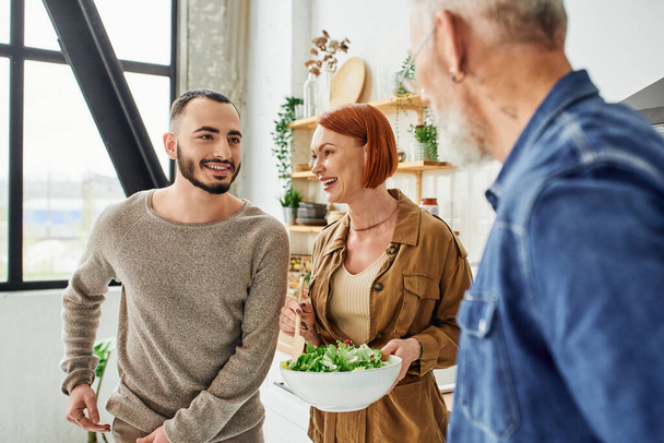 happy redhead woman holding bowl with fresh salad near son and blurred husband in kitchen - Foto, Imagem