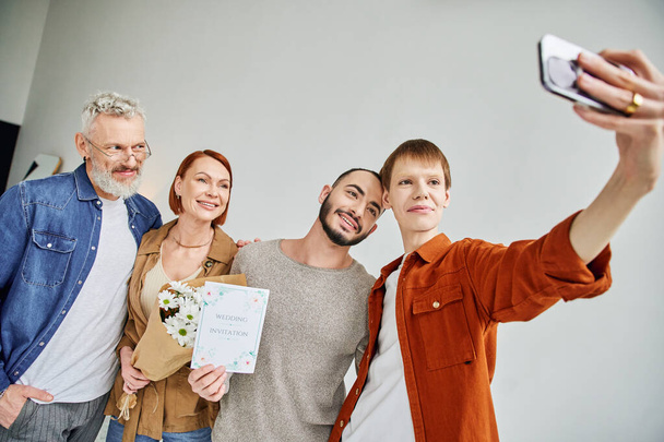 onnellinen homo mies ottaa selfie poikaystävän ja vanhempien kanssa, joilla on kukkia ja häät kutsukortti - Valokuva, kuva