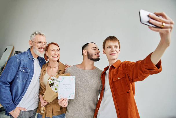 Rotschopf-Mann macht Selfie mit homosexuellem Partner und glücklichen Eltern mit Heiratseinladung und Blumen - Foto, Bild