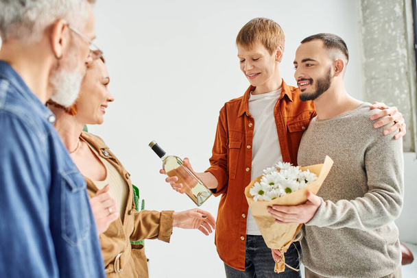 cheerful gay couple holding flowers and wine bottle near parents at home - Фото, зображення