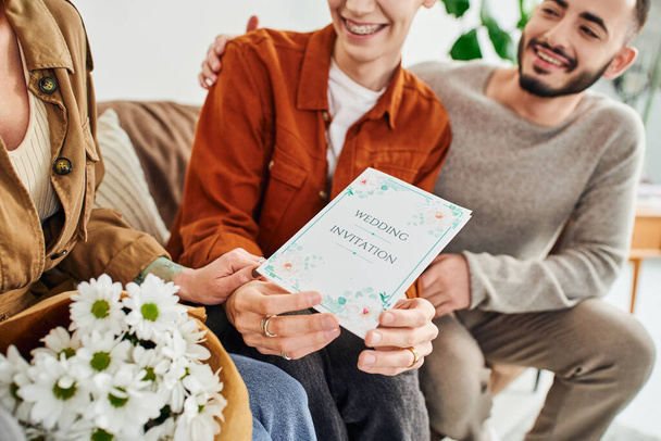 happy gay man holding wedding invitation near boyfriend and mother with flowers at home - Photo, Image