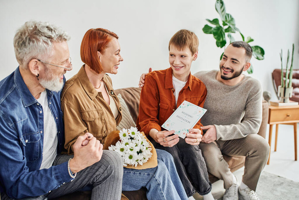pleased gay man showing wedding invitation to parents sitting on couch with flowers - Foto, Bild