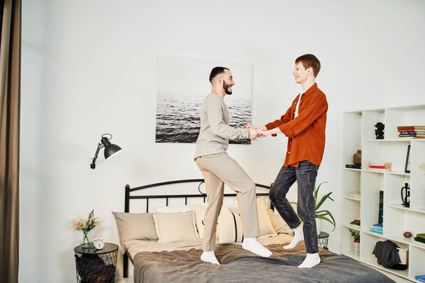 full length of young gay couple holding hands and having fun on bed in modern bedroom - Foto, afbeelding