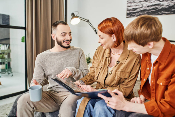  bearded gay man talking to redhead woman looking at family photo album at home - Photo, image