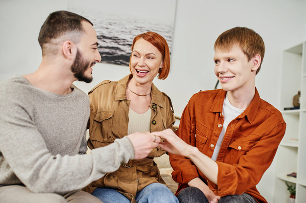 emocionado mujer riendo cerca gay hombre e hijo mostrando boda anillo en casa - Foto, imagen