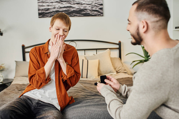 opgewonden roodharige man bedekking mond met handen in de buurt wazig vriendje maken huwelijksaanzoek in slaapkamer - Foto, afbeelding