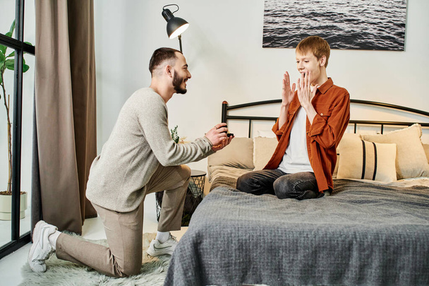 изумленный гей показывает вау жест рядом с бойфрендом делая предложение о браке в спальне - Фото, изображение