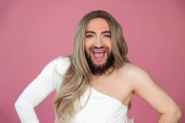Äußerst glücklicher Transgender mit blonder Perücke und weißem Kleid hält die Hände auf den Hüften und lacht laut, posiert isoliert vor rosa Hintergrund - Foto, Bild