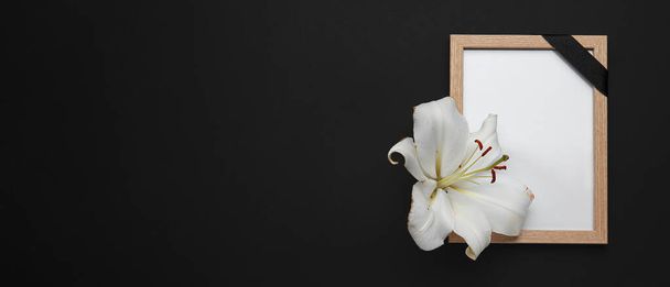Photo frame with black funeral ribbon and lily flower on dark background with space for text - Photo, Image