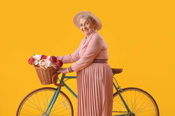 Mulher sênior com bicicleta e cesta de flores no fundo amarelo - Foto, Imagem