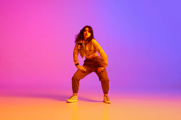 Active, artistic young woman, hip hop dancer in sport style clothes dancing against gradient pink purple background in neon light. Concept of contemporary dance, youth, hobby, action and motion - Photo, image