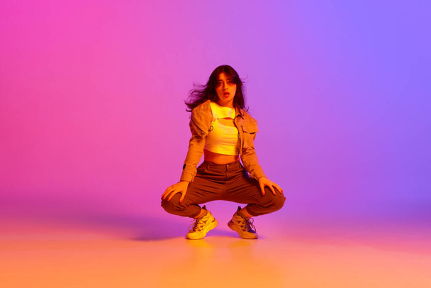Young expressive girl in sport style clothes dancing modern dance against gradient pink purple background in neon light. Concept of contemporary dance, youth, hobby, action and motion - Photo, image