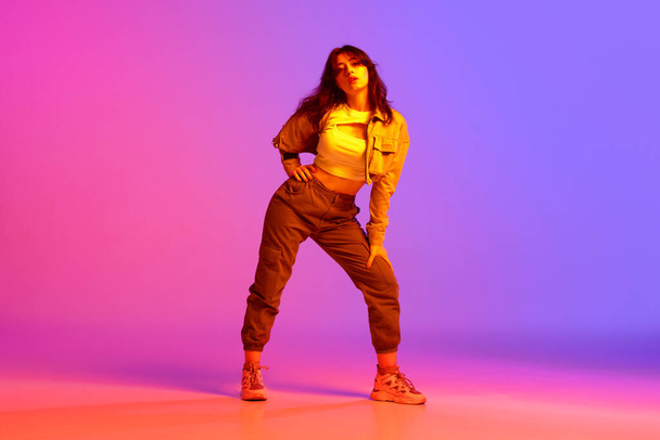 Artistic, beautiful, young girl dancing hip-hop against gradient pink purple background in neon light. Model in casual clothes. Concept of contemporary dance, youth, hobby, action and motion - 写真・画像