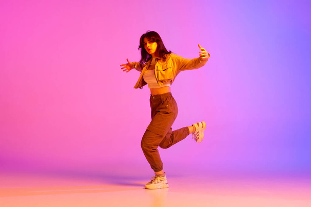 Immagine dinamica di una giovane ballerina in abiti sportivi che danza sullo sfondo viola rosa sfumato in luce al neon. Concetto di danza contemporanea, gioventù, hobby, azione e movimento - Foto, immagini