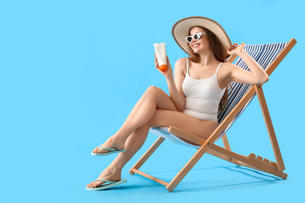 Beautiful young woman with sunscreen cream sitting on deck chair against blue background - Foto, Imagen