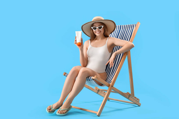 Beautiful young woman with sunscreen cream sitting on deck chair against blue background - Фото, изображение