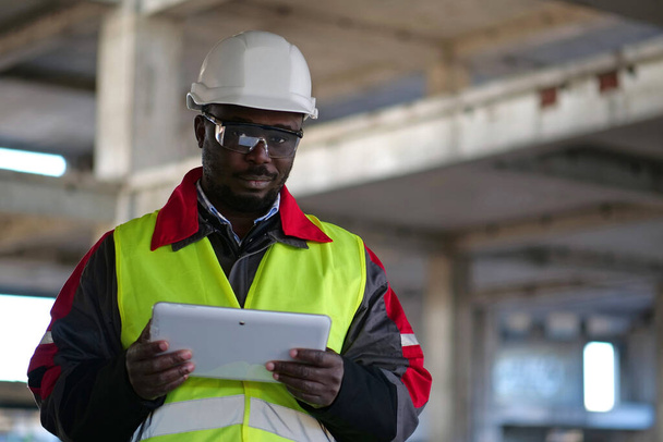 Afrikkalainen amerikkalainen työntekijä kypärässä ja suojalasit seisoo rakennustyömaalla, pitää käsissä tabletin tietokone, katsoo kameraan ja hymyilee - Valokuva, kuva
