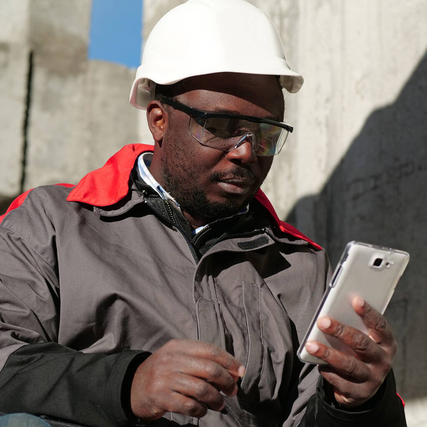 Trabajador afroamericano en sombrero duro y gafas protectoras con teléfono inteligente se sienta en el sitio de construcción. Workman sostiene en las manos el teléfono inteligente blanco - Foto, Imagen